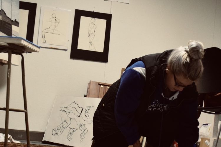 Woman wearing sweatshirt and glasses kneels over large paper inside art studio